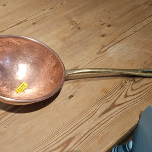 May include: A hammered copper cooking pan with a brass handle. The pan has a round, shallow bowl shape and a smooth, shiny surface.