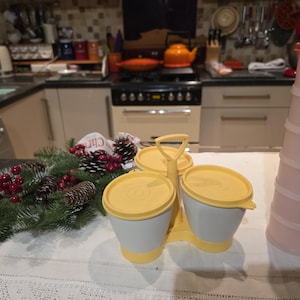 Puede incluir: Un juego de tres recipientes para alimentos blancos y amarillos con tapas amarillas y un asa. Una pila de recipientes rosas está a la derecha. Una guirnalda navideña con piñas y bayas rojas está en primer plano.