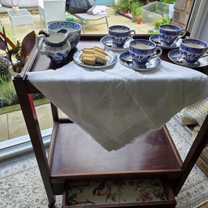 May include: A dark wooden tea trolley with a white cloth, displaying blue and white china teacups, saucers, a milk jug, and a bowl. Small cakes are on a plate. The trolley is near a window overlooking a garden.