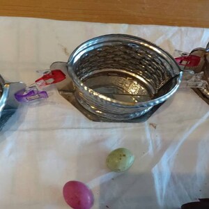 May include: Three silver metal baskets with a woven design are connected together with clips. The baskets are on a white cloth surface. Two colorful candy eggs are in the foreground.
