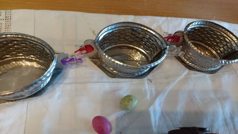 May include: Three silver metal baskets with a woven design are connected together with clips. The baskets are on a white cloth surface. Two colorful candy eggs are in the foreground.