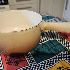 May include: A brown and beige ceramic fondue pot with a handle. The pot is sitting on a colourful checkered tablecloth.