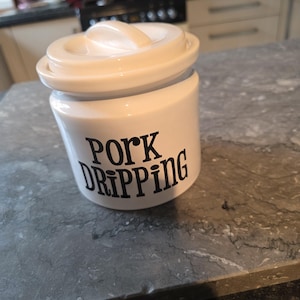 以下が含まれることがあります： クリーム色のセラミックジャーで、蓋とハンドルが付いており、黒い大文字で「PORK DRIPPING」とラベル付けされています。円筒形のジャーは灰色のカウンタートップに置かれています。蓋には小さなハンドルが付いています。