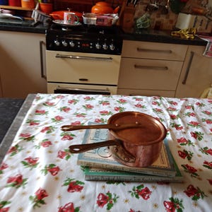 May include: A copper saucepan with a lid rests on a stack of books on a floral tablecloth. The pan has two handles and a matching copper lid. The tablecloth has a pattern of red roses and green leaves. The kitchen in the background has a stove and cabinets.