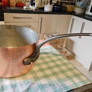 May include: A large copper pot with a long, curved handle. The pot is sitting on a checkered tablecloth.