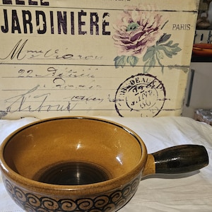Puede incluir: Una olla de fondue de cerámica marrón con un mango de madera marrón oscuro. La olla tiene un patrón de espiral decorativo alrededor del borde.