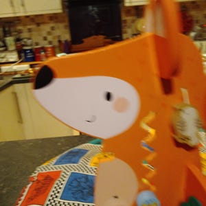 May include: An orange fox-shaped decoration with white and black facial features. The fox has a light pink cheek and a small smile. The decoration is on a patterned surface.