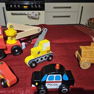 May include: A set of five wooden toy vehicles, including a red tow truck with a yellow crane, a yellow construction vehicle, a brown tractor, a red race car, and a black police car. The toys are all made of wood and have painted details.