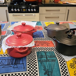 May include: Four small ceramic casserole dishes with lids. Two are red, one is grey, and one is black. The dishes are on a colourful tablecloth with a black and white checkered pattern.