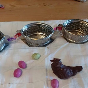 May include: Four silver metal baskets with handles, a chocolate Easter bunny, and pink and green Easter eggs on a white tablecloth.