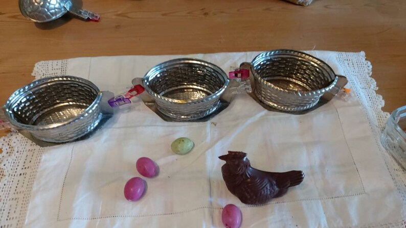 May include: Four silver metal baskets with handles, a chocolate Easter bunny, and pink and green Easter eggs on a white tablecloth.
