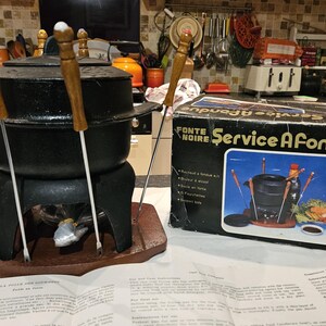 May include: A black fondue set with wooden handles and metal forks, alongside its original box. The box displays an image of the fondue set and the words "Service A Fondue". The set is on a wooden base.