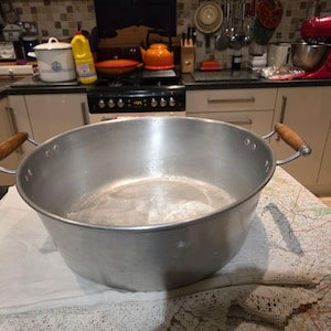 May include: Large, aluminium cooking pot with wooden handles. The pot is round with a wide opening and sits on a white cloth with a map design. The handles are attached to the sides with metal rivets.