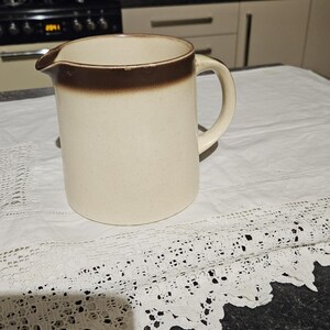 May include: A brown and white ceramic pitcher with a brown rim and a handle. The pitcher is sitting on a white lace tablecloth.
