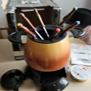 May include: A brown fondue pot with a black base and six fondue forks. The pot is filled with melted chocolate and the forks are arranged in a circle around the pot. There is a small black ladle and a container of fondue chocolate on the table.