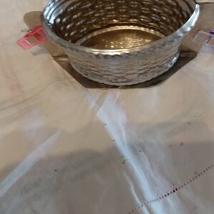 May include: A silver metal container with a woven pattern. The container is empty and sitting on a white tablecloth.