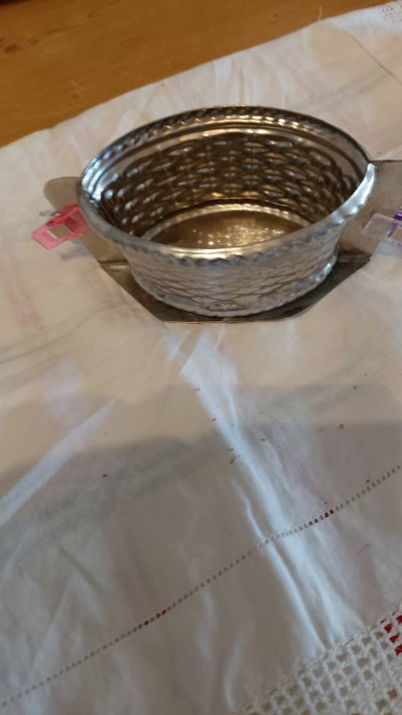 May include: A silver metal container with a woven pattern. The container is empty and sitting on a white tablecloth.