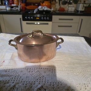 May include: A copper saucepan with a lid and two side handles. The pot is on a white, lace-trimmed tablecloth. The lid handle is gold-coloured. The background shows a kitchen with a stove and cabinets.