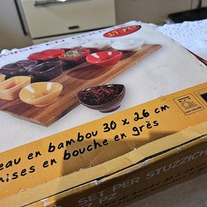 Op de afbeelding: Een set van 12 kleine keramische kommen met een bamboe serveerblad. De kommen zijn in verschillende kleuren, waaronder rood, geel en wit. Het blad is 30 cm bij 26 cm. De tekst op de doos luidt "Plateau en bambou 30 x 26 cm, 12 mises en bouche en grès, Set per Stuzzichini".