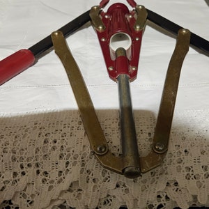 May include: A red and gold bottle opener with black handles. The opener is on a white tablecloth with lace trim.