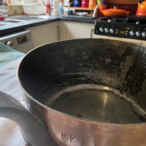 May include: A large copper cooking pot with a dark interior and a long, curved handle. The pot has a shiny, reddish-brown exterior and is marked with the initials "FF". This vintage kitchenware item is likely used for cooking and is a classic piece.