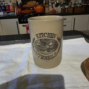 May include: A beige ceramic kitchen utensil holder with black text that reads "KITCHEN UTENSILS" around a graphic of crossed forks and spoons. The cylindrical holder is on a white cloth.