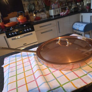 May include: A copper-colored frying pan with a long handle and a clear glass lid. The pan is sitting on a colorful checkered kitchen towel. The kitchen in the background has white cabinets and a black stove.
