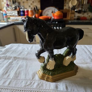 May include: A black horse figurine with white markings on its legs and face, posed on a green and tan base. The horse is sculpted in a dynamic, prancing stance, showcasing its muscular form. This decorative piece is likely ceramic or porcelain.