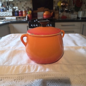 May include: A round, bright orange ceramic pot with two side handles and a matching lid. The lid features a dark brown, circular knob handle at the top. The pot has a smooth, glossy finish and a slightly bulbous shape, resting on a flat base.