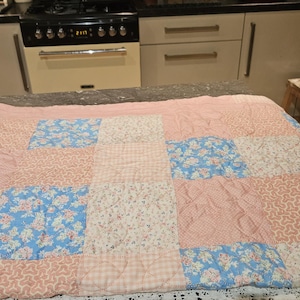 May include: A patchwork quilt with a pink, blue and white floral pattern. The quilt is made of different fabrics and has a quilted texture.