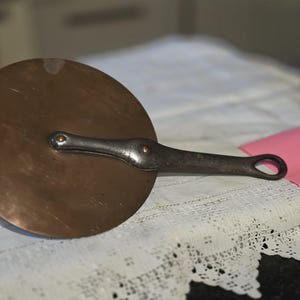 May include: A circular copper pan lid with a riveted, silver-toned handle. The handle has a loop at the end. The lid sits on a white, lace-trimmed surface, with a pink square of paper nearby. Kitchenware.