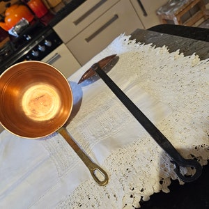 May include: A copper ladle with a long handle sits on a white lace tablecloth next to a black metal ladle with a loop handle.