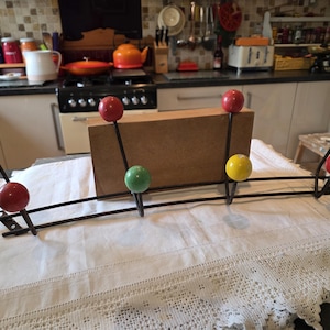 May include: A retro coat rack with a black metal frame and six coloured wooden ball knobs. The knobs are red, yellow, and green. The rack is displayed on a white tablecloth, suggesting a vintage or mid-century modern design.