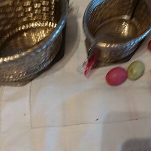 May include: Three silver metal bowls with a woven pattern, sitting on a white tablecloth with a lace trim. There are several pink and green candy eggs scattered around the bowls.