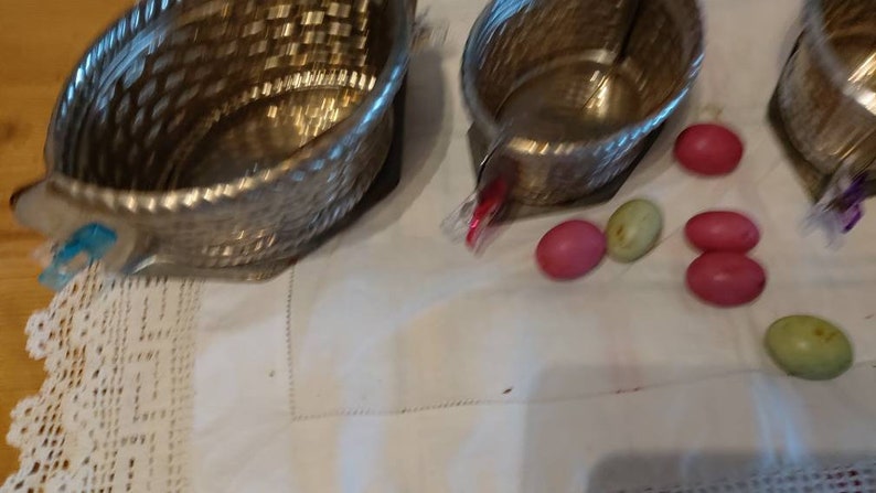 May include: Three silver metal bowls with a woven pattern, sitting on a white tablecloth with a lace trim. There are several pink and green candy eggs scattered around the bowls.