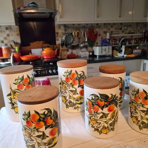 May include: Six white ceramic kitchen canisters with wooden lids. Each features a design of oranges and green leaves. The cylindrical canisters are of varying sizes, suitable for storing dry goods.
