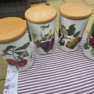 May include: Four cylindrical ceramic canisters with light-coloured wooden lids. Each white canister features a detailed botanical illustration of different fruits, including red apples, purple figs, and green pears, with green leaves and small blossoms.
