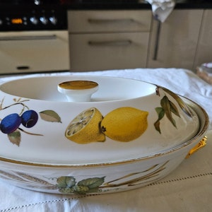 May include: A white ceramic serving dish with a gold rim and a lid. The dish features a floral design with lemons, plums, and leaves. The lid has a knob with a gold top.