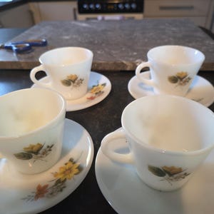May include: Four white teacups and saucers with a floral design. Each cup has a handle and sits on a matching saucer. The floral design features yellow and orange flowers with green leaves. The cups and saucers are arranged on a dark surface.