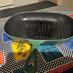 May include: A green cast iron grill pan with a black interior and raised grill lines. The pan is oval shaped and has a single handle.