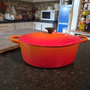 May include: A round enameled cast iron Dutch oven featuring a vibrant gradient finish that transitions from a deep red at the base to a bright orange on the lid. The pot includes two side handles and a central black knob on the matching lid.