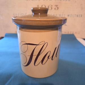 Könnte beinhalten: Ein zylindrischer Keramikbehälter mit hellbeigem Körper und dunkelbraunem Deckel. Das Wort Flour ist in dunkelbrauner Schreibschrift auf der Vorderseite aufgedruckt. Der Behälter hat eine glatte, glänzende Oberfläche und einen passenden braunen Knauf.