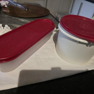Peut inclure: Deux récipients de stockage alimentaires en plastique blanc avec des couvercles rouges. L'un est rectangulaire, l'autre est rond. Les deux récipients sont sur une surface blanche. Les couvercles sont rouge foncé.