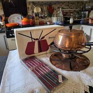 May include: A vintage copper fondue pot with a black handle and stand, and a box with a red and white graphic, plus a set of fondue forks. The fondue pot is approximately 20 cm in diameter.