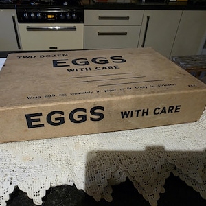 Puede incluir: Una caja de huevos vintage de cartón marrón con texto negro "TWO DOZEN EGGS WITH CARE". La caja está sobre un mantel de encaje blanco. También se ve el texto "Wrap each egg separately in paper".