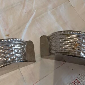 May include: Two silver metal molds shaped like baskets. The molds are half-circle shaped and have a woven pattern.