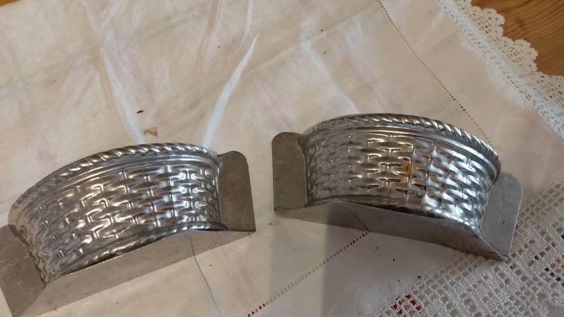 May include: Two silver metal molds shaped like baskets. The molds are half-circle shaped and have a woven pattern.