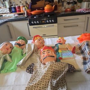 May include: A collection of vintage hand puppets with painted faces and fabric clothing. The puppets have various expressions and wear hats and outfits in shades of green, red, orange, and brown. They are arranged on a white surface.