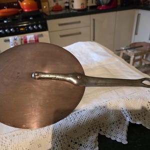 May include: A round copper pot lid with a long, silver-coloured handle. The lid has a slightly domed shape. The handle is riveted and has a loop. The lid's diameter is approximately 25 cm. Kitchenware.