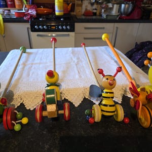 May include: A selection of brightly coloured wooden pull-along toys, including a duck and a bee. Each toy has a long wooden handle and is painted with vibrant colours and details. The toys are displayed on a table, with a white tablecloth.